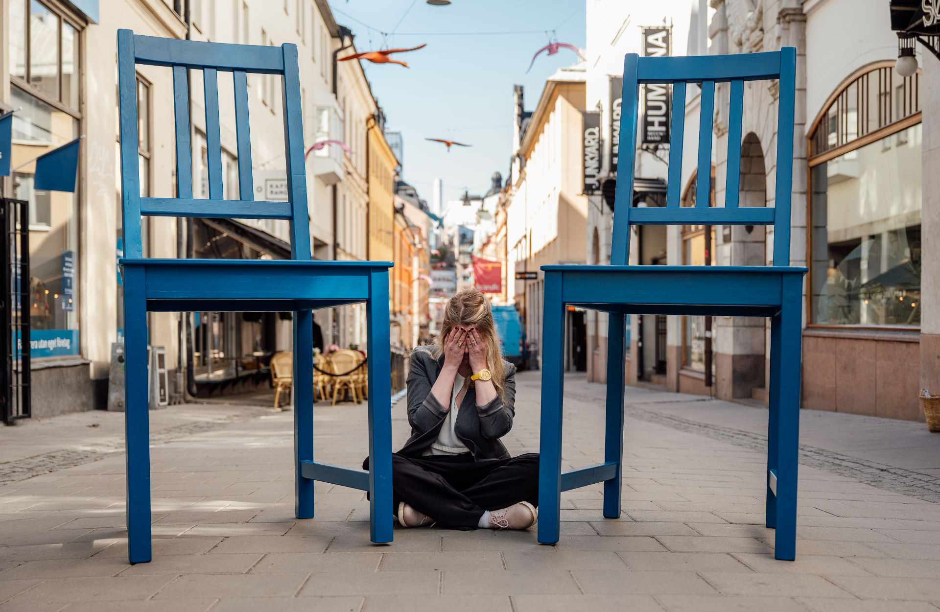 En person sitter med ansiktet i händerna mellan två överdimensionerade blå stolar på en gata i Stockholm.